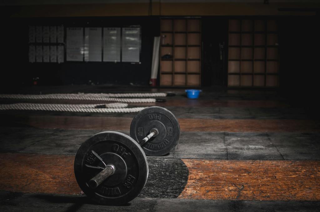 A pair of black dumbbells resting on a gym floor with a rustic, dimly lit background featuring wooden panels and gym equipment.